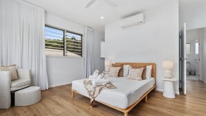 A modern bedroom with a wooden bed, a sitting area with a chair, a bedside table with a lamp, and a large window with curtains. - Beachmont Bliss Palm Cove 3 Bedroom Holiday Home (Palm Cove)