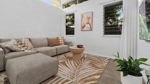 A modern living room with a grey sectional sofa, a round coffee table, and a large window with blinds. There is a painting on the wall and a potted plant on the right. - Beachmont Bliss Palm Cove 3 Bedroom Holiday Home (Palm Cove)