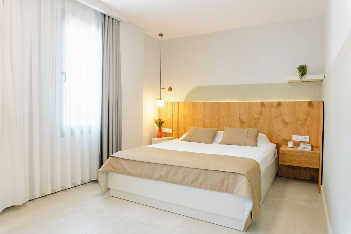 A modern hotel room with a large bed, a bedside table with a lamp, a nightstand with a phone, and a small plant on the nightstand.