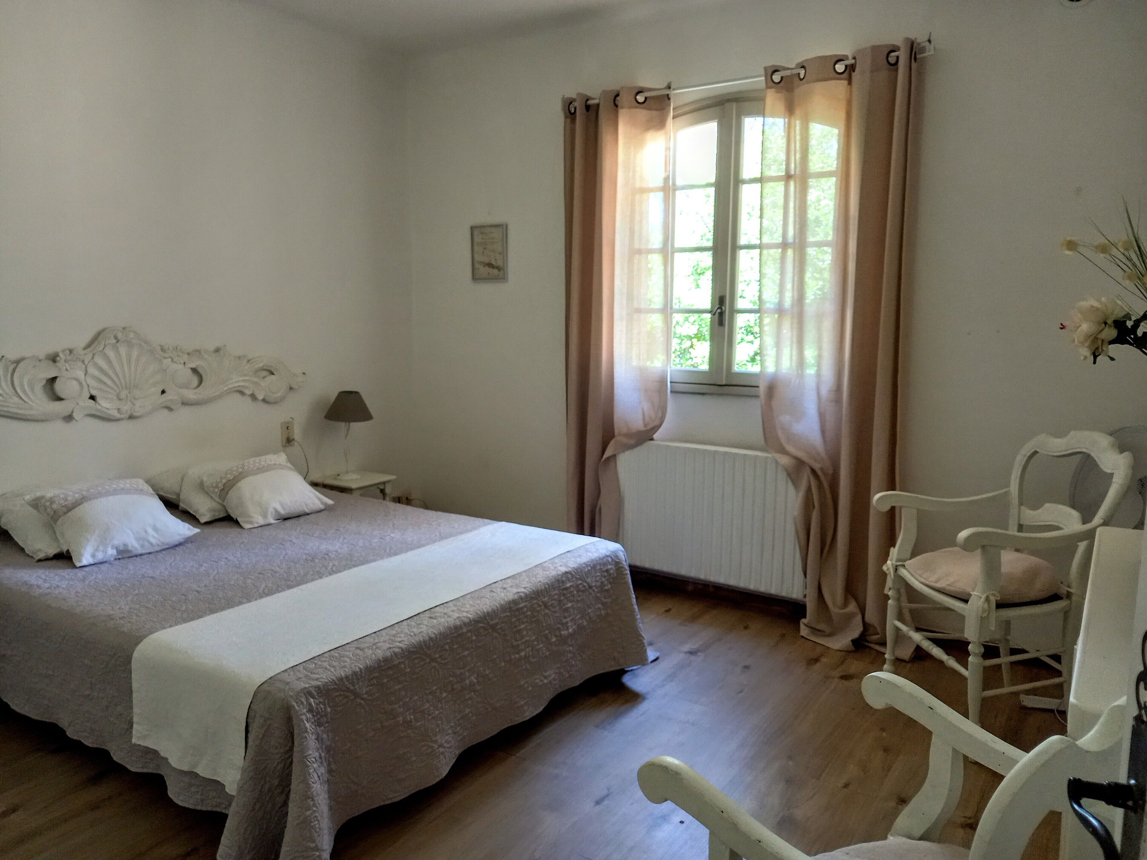 A bedroom with a bed, a chair, a nightstand, a lamp, and a window with curtains.
