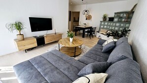 A modern living room with a grey sectional sofa, a wooden coffee table, a flat-screen TV, a dining area with a table and chairs, and a decorative cabinet with plants. - Apartment Marina (Oberzent)