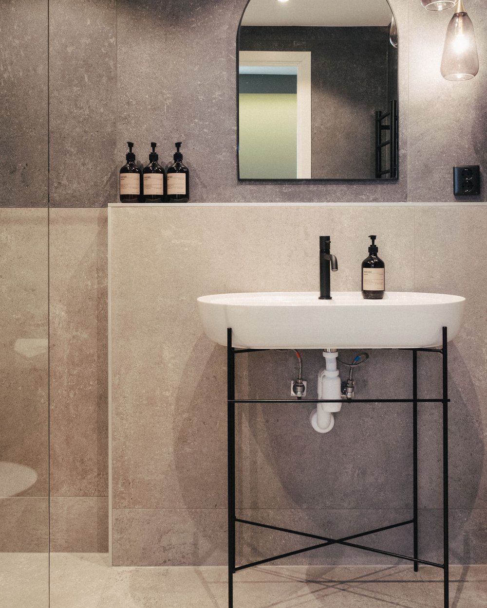 A modern bathroom with a white sink, black faucet, and a mirror above it. There are two bottles of soap on the wall and a light fixture above the mirror.