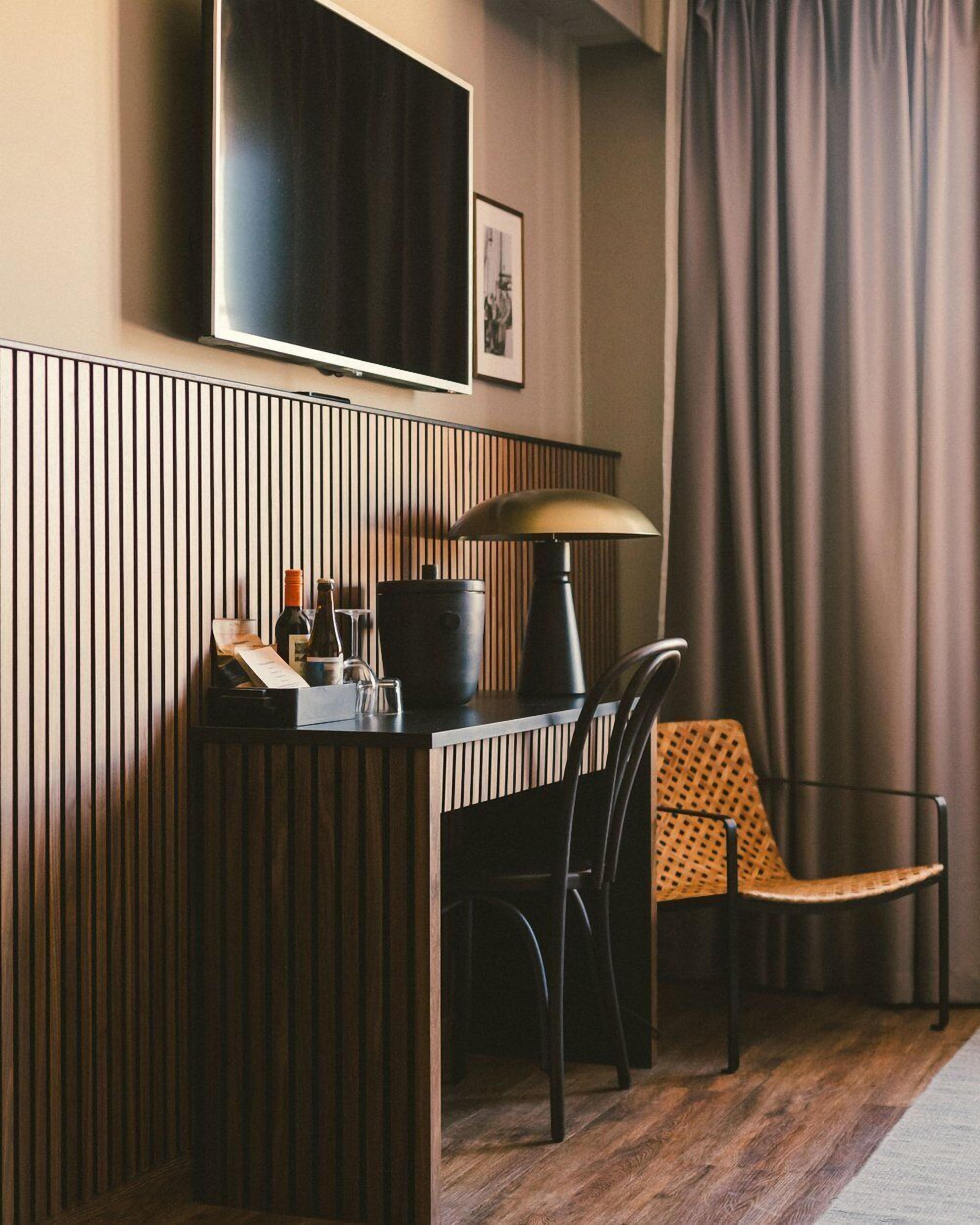 A hotel room with a wooden panel wall, a flat-screen TV mounted above a desk, a lamp, a chair, and a bed with a patterned cover.