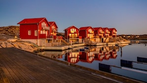 A row of red wooden buildings with balconies, a wooden deck, and a calm body of water reflecting the buildings. - Hotel Strana (Hälleviksstrand)