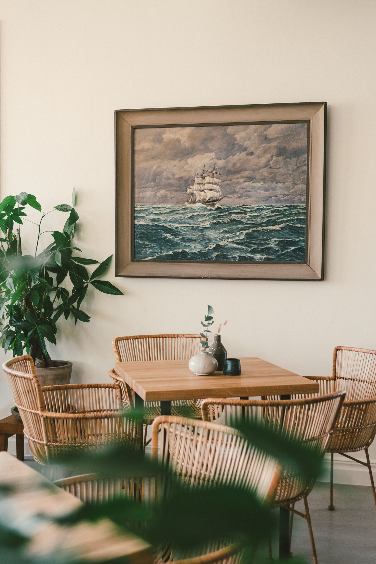 A dining area with a wooden table and wicker chairs, a framed painting of a ship on the sea, and a large potted plant.