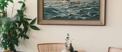 A dining area with a wooden table and wicker chairs, a framed painting of a ship on the sea, and a large potted plant.