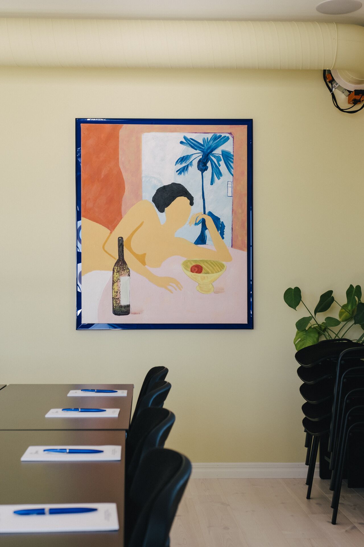 A framed painting of a person reclining with a bottle and a bowl on a table, a ceiling vent, and a stack of black chairs.