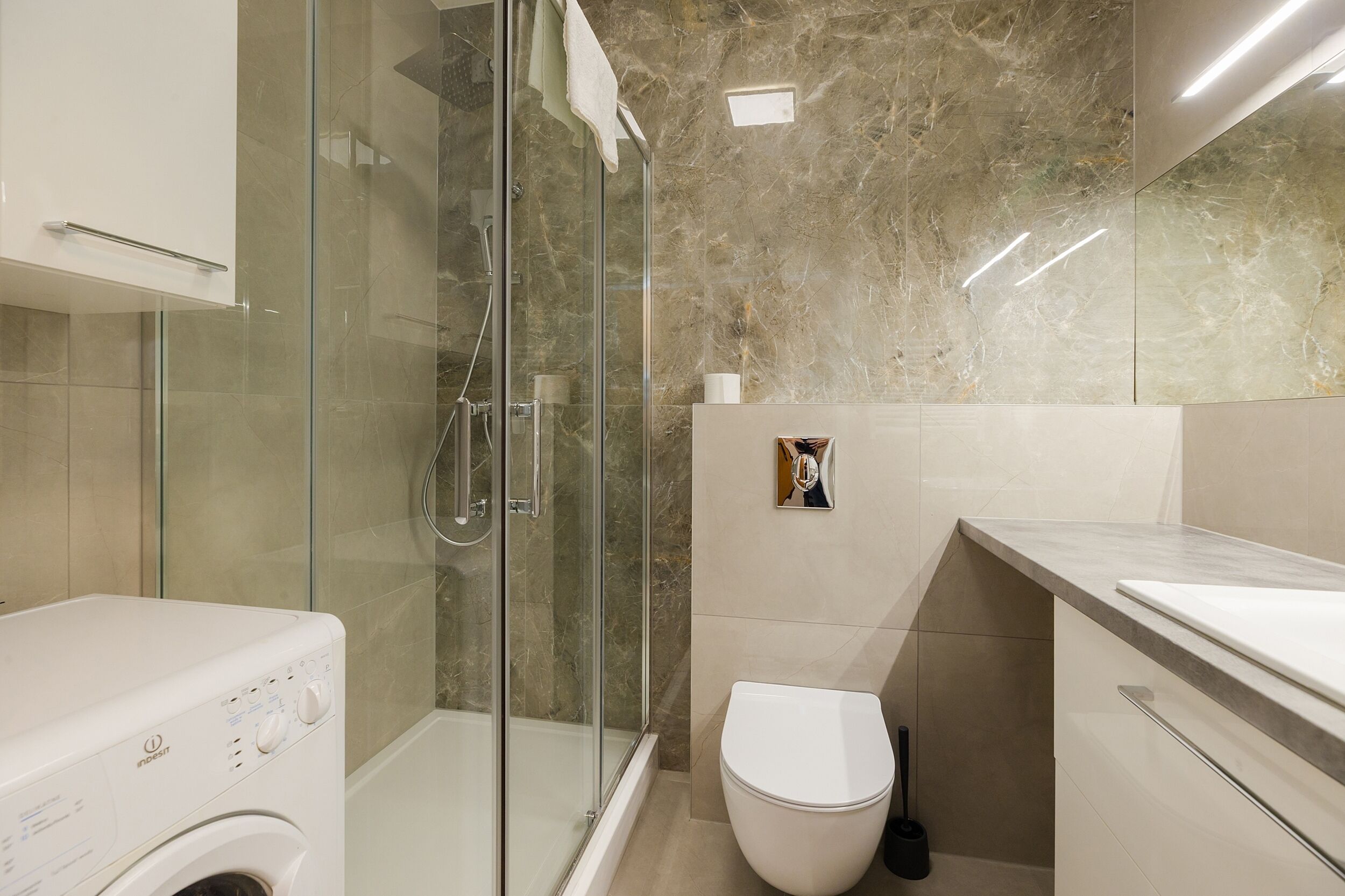 Een moderne badkamer met een douchecabine van glas, een witte wasmachine, een toilet en een achterwand van marmer.