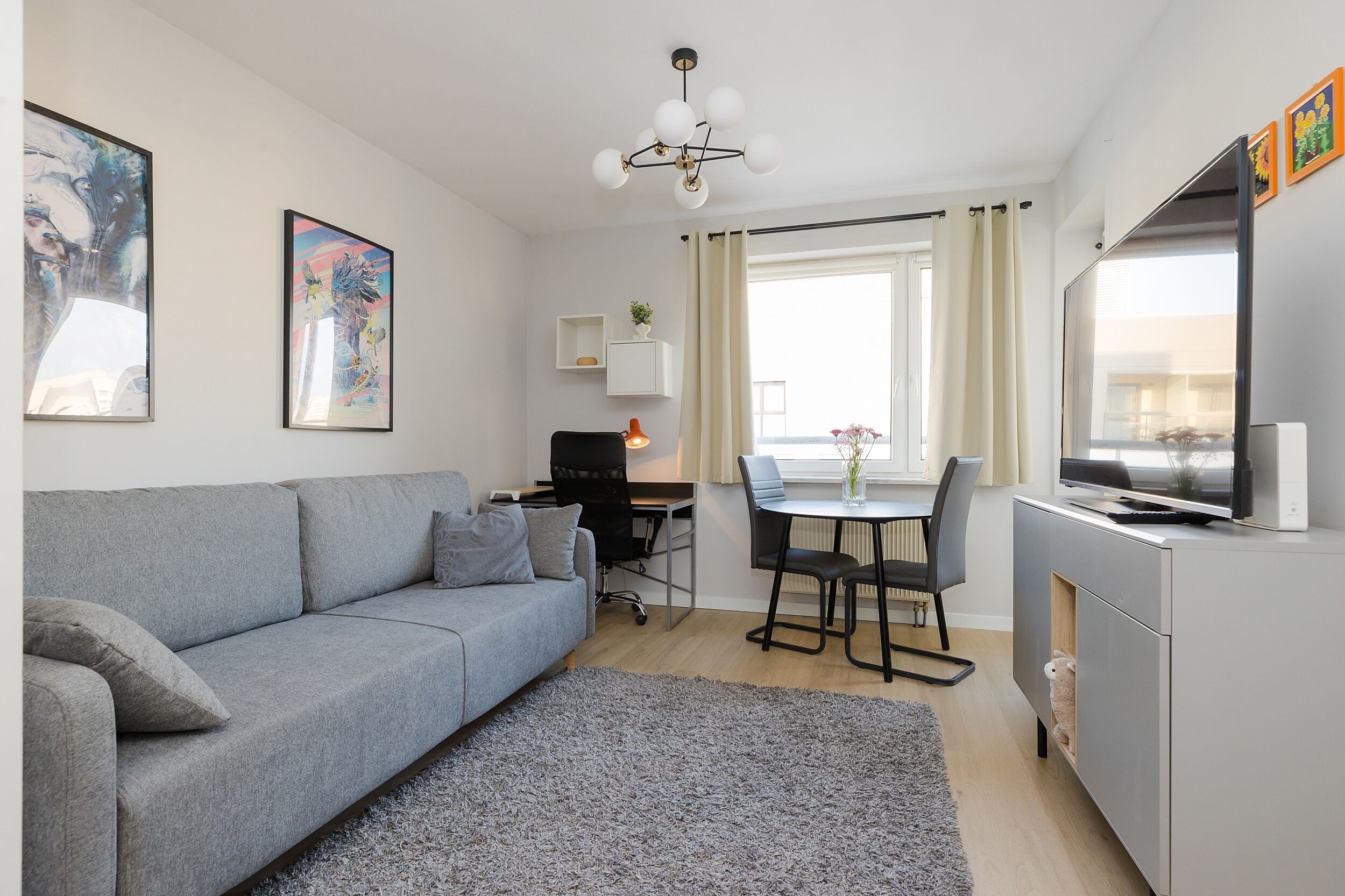 Un salon moderne avec un canapé gris, une table à manger avec des chaises, un petit bureau et un téléviseur posé sur un meuble.
