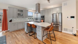A modern kitchen with grey cabinets, a central island with stools, stainless steel appliances, and a large window. - Spacious Denver Home with Hot tub and Rooftop (Denver)