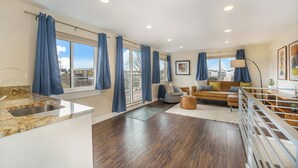 A modern kitchen with a granite countertop and stainless steel sink, a wooden dining table with chairs, a sofa, and a coffee table in a living area with large windows and blue curtains. - Spacious Denver Home with Hot tub and Rooftop (Denver)