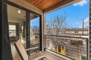 Een balkon met uitzicht op een boom en gebouwen, een stoel en een tafel met een lamp.