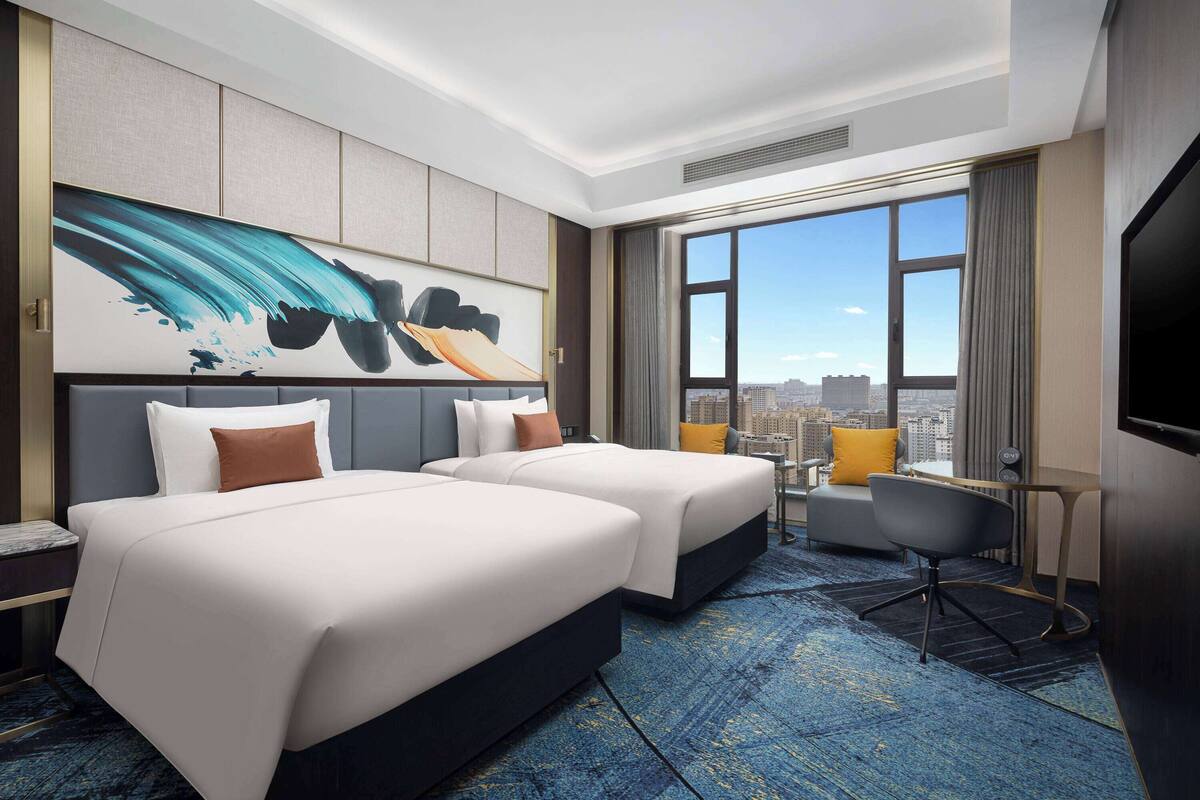 A hotel room with two beds, a desk, a chair, and a large window with a city view.