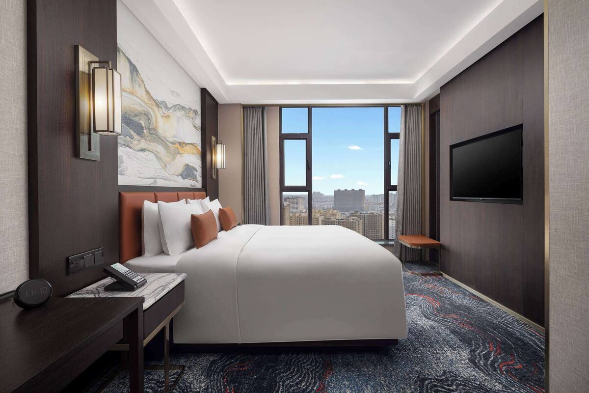 A hotel room with a large bed, a desk with a telephone, a flat-screen TV, and a city view through large windows.