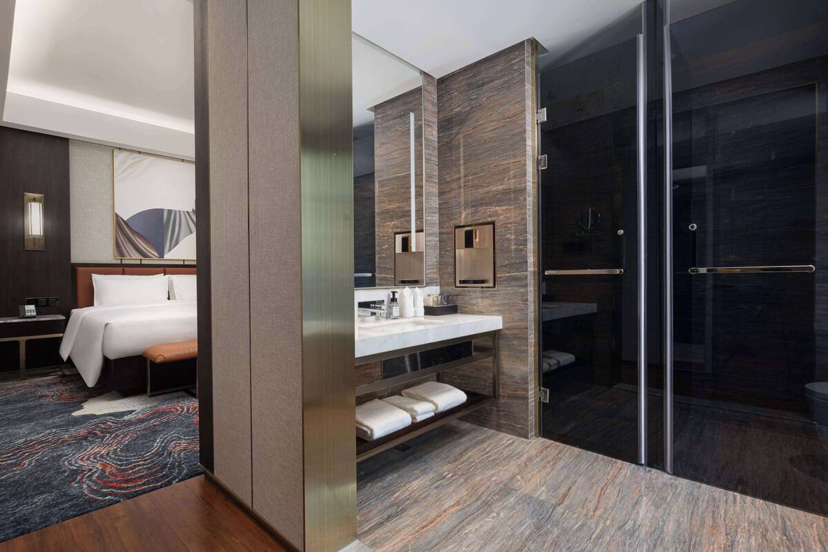 A modern hotel room with a large bed, a bathroom with a glass shower, and a marble floor.