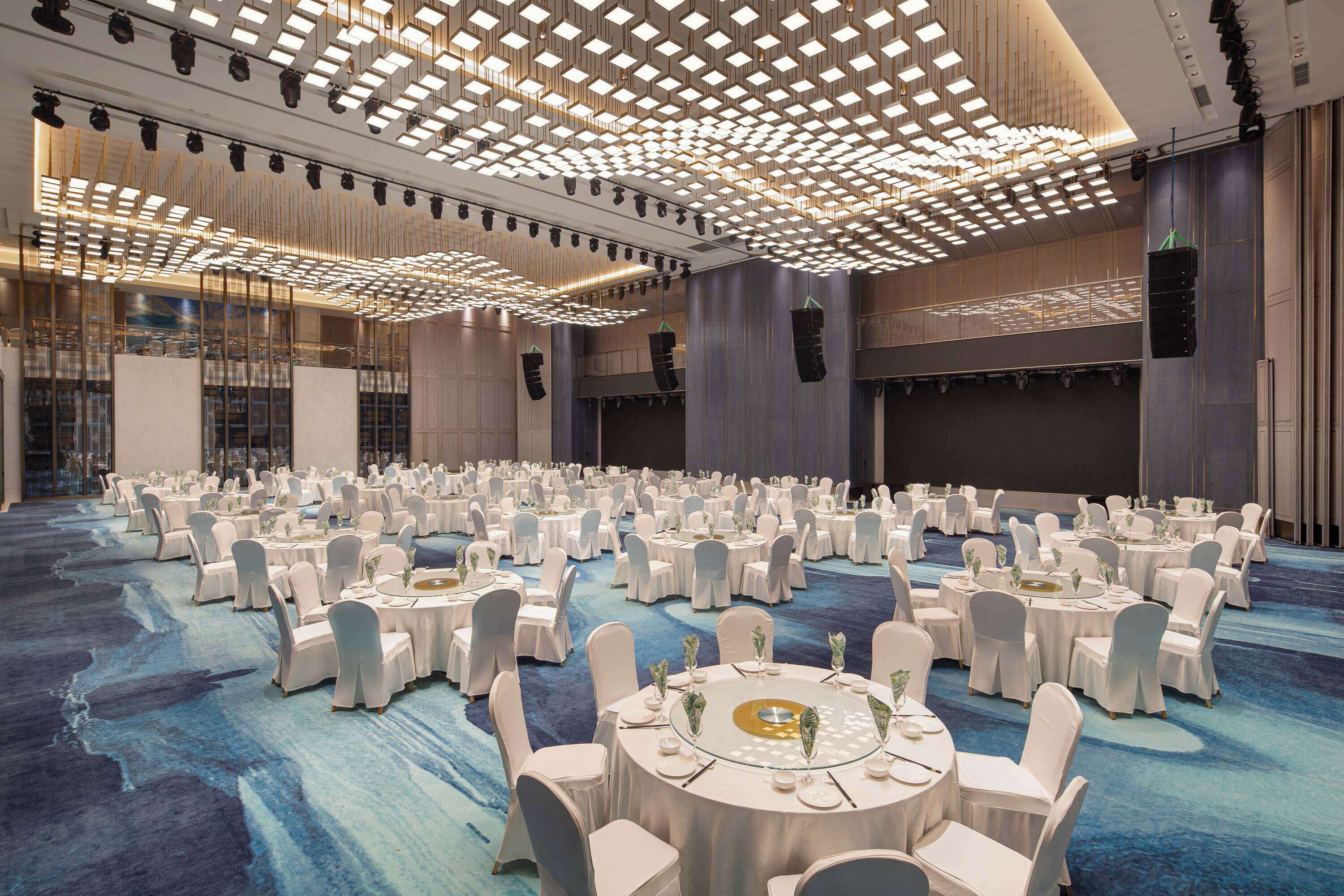 Un amplio salón de banquetes con mesas redondas, sillas blancas y una alfombra azul.