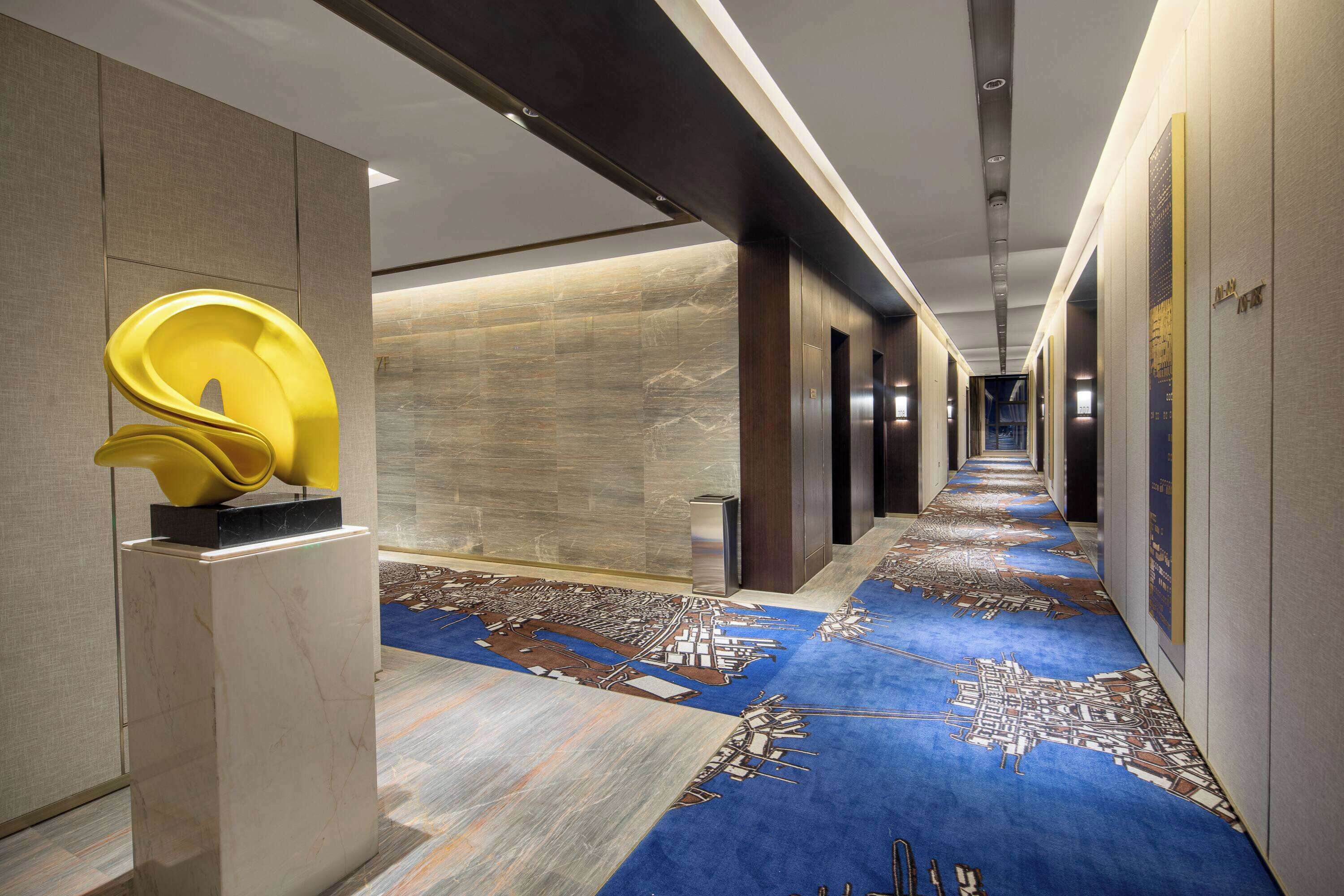 A modern hotel corridor with a blue patterned carpet, a yellow sculpture, and a cityscape design on the floor.