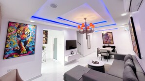 A modern living room with a sofa, a coffee table, and a TV. There is a painting on the wall and a chandelier. - The Muse By LVG (Lekki)