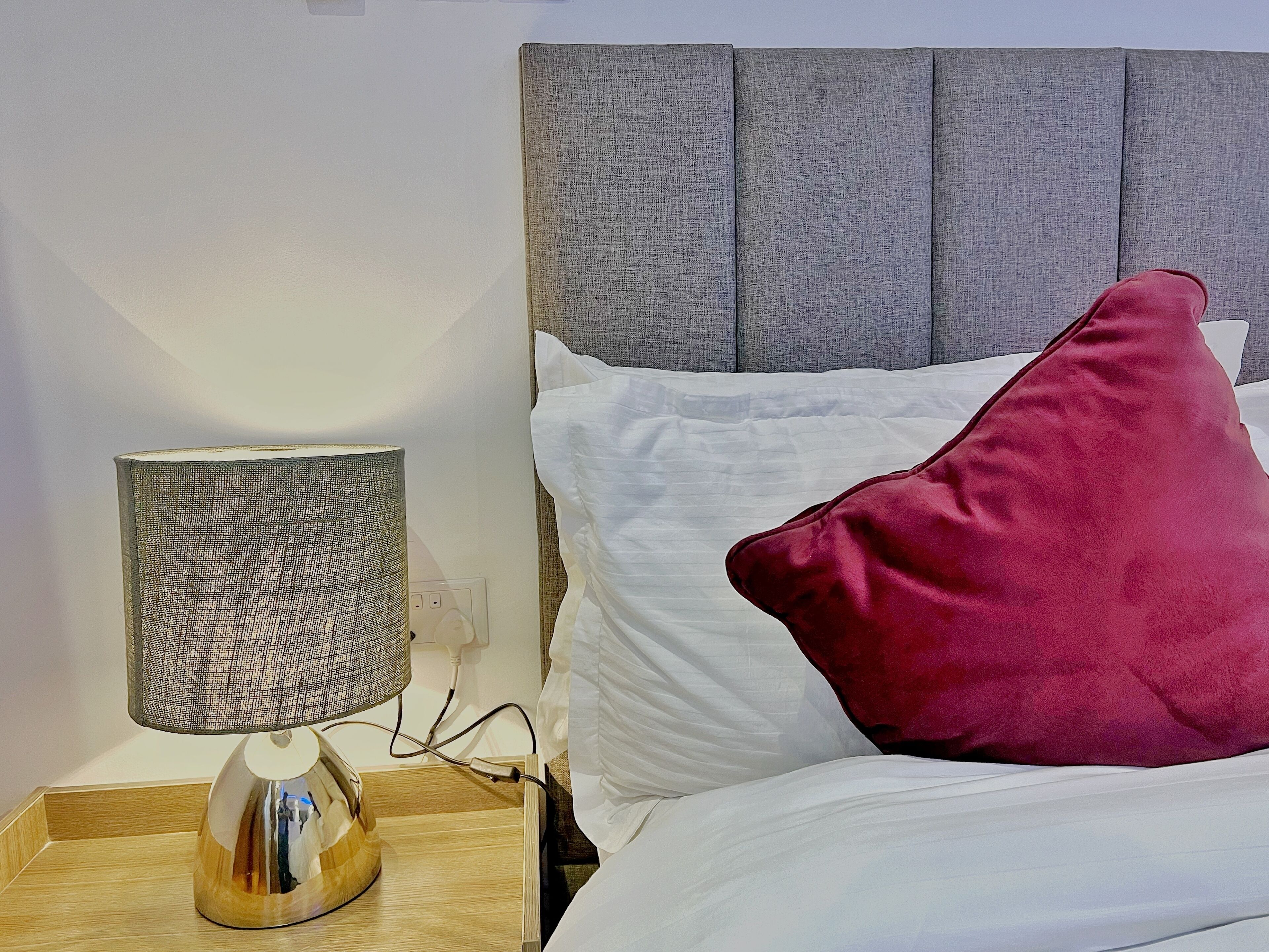 Een net opgemaakt bed met witte en rode kussens, een grijze hoofdbord en een nachtkastje met een lamp en oplader.