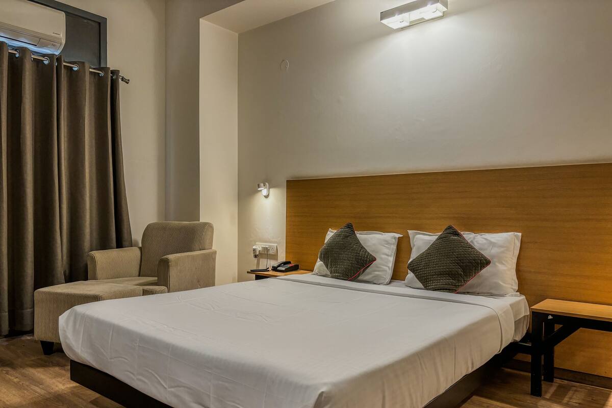 A hotel room with a large bed, a chair, a bedside table with a lamp, and a wall-mounted air conditioning unit.