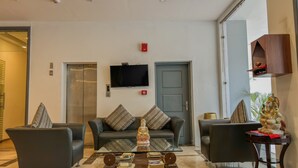 A modern living room with a sofa set, a coffee table with books and decorative items, a television mounted on the wall, and a door with a security system. - Hotel Bizzotel (Gurugram)