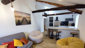 Un salon moderne comprenant un canapé, deux fauteuils poire, une table basse en bois et un coin cuisine avec un tabouret de bar.