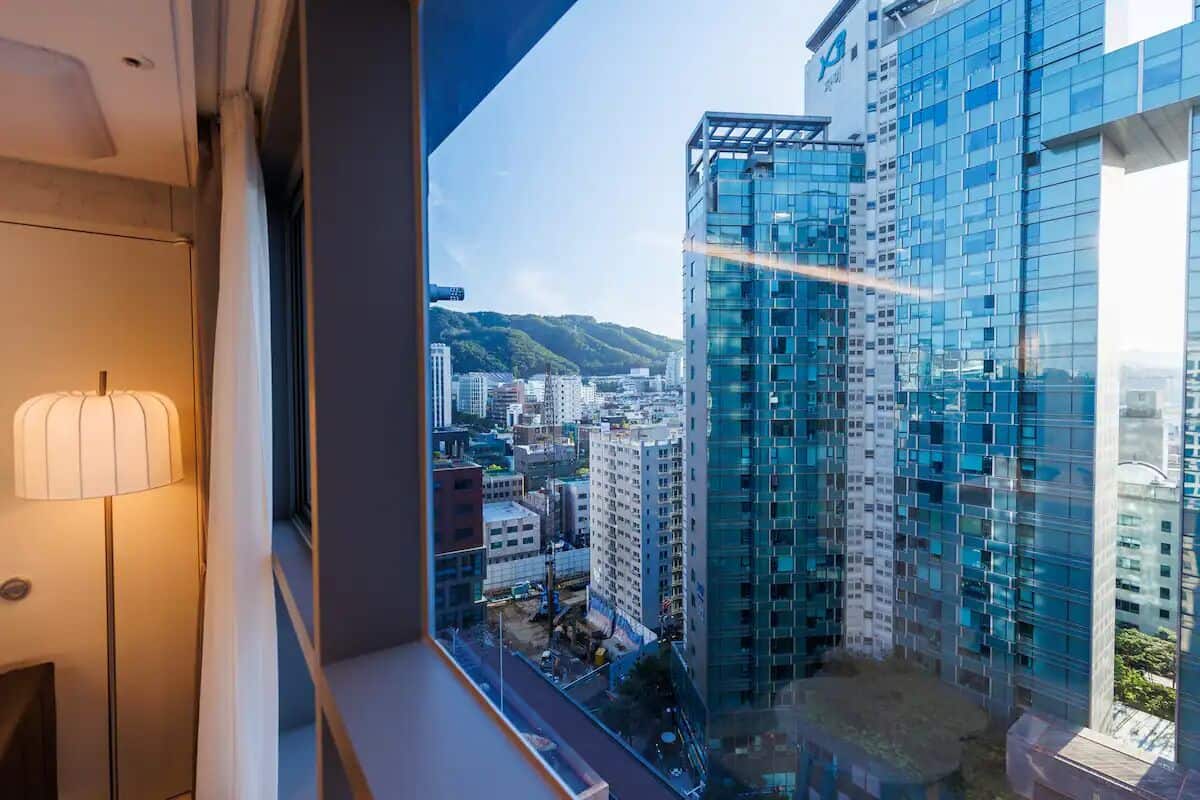 A high-rise building with a modern glass facade, viewed from a window with a lamp on the left.