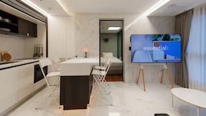 Habitación de hotel moderna con cocina, una mesa de comedor con sillas, cama y televisor sobre un soporte.