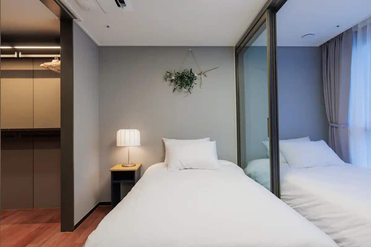 A modern hotel room with a large bed, a bedside table with a lamp, a wall-mounted plant, and a sliding glass door leading to a balcony.