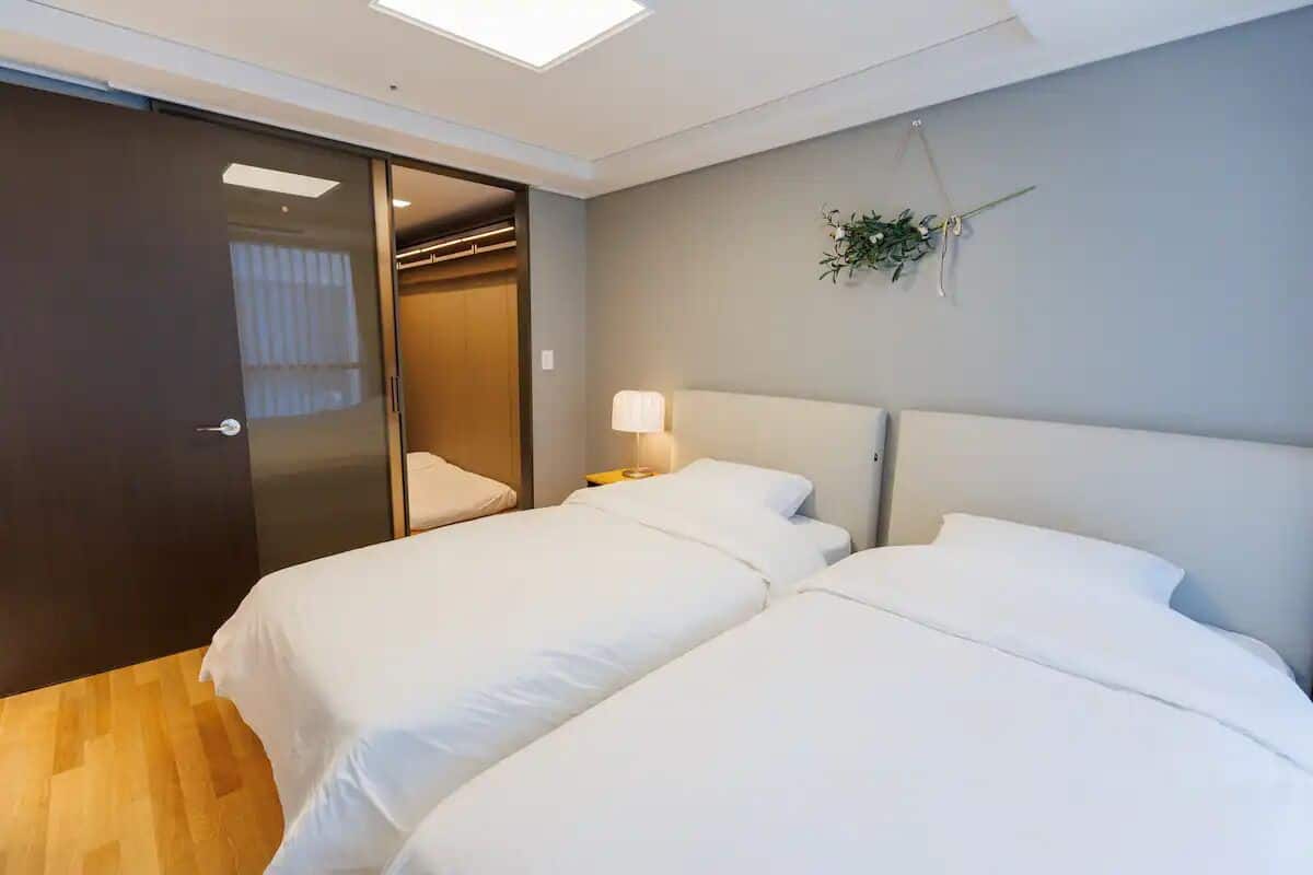 A hotel room with two beds, a wooden floor, a sliding door, a bedside lamp, and a wall-mounted plant.