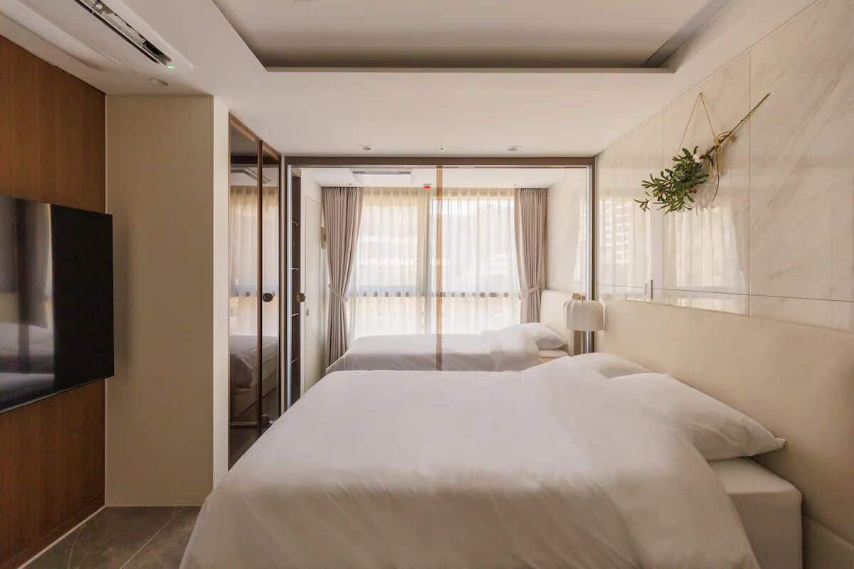 A hotel room with two beds, a TV, and a view of the city through large windows.