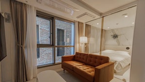 Una habitación de hotel moderna con un sofá de cuero, una mesita, una cama con ropa de cama blanca y vistas a un edificio de ladrillo a través de grandes ventanales.