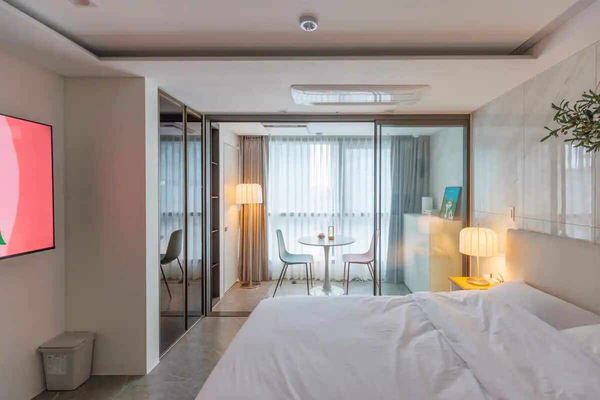 A modern hotel room with a large bed, a TV, a balcony with a table and chairs, and a view of the city.