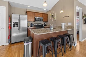 A kitchen with a stainless steel refrigerator, a microwave, a stove, and a granite countertop. There are four black barstools and a trash can. - Carriage Home with early check in (Indianapolis)