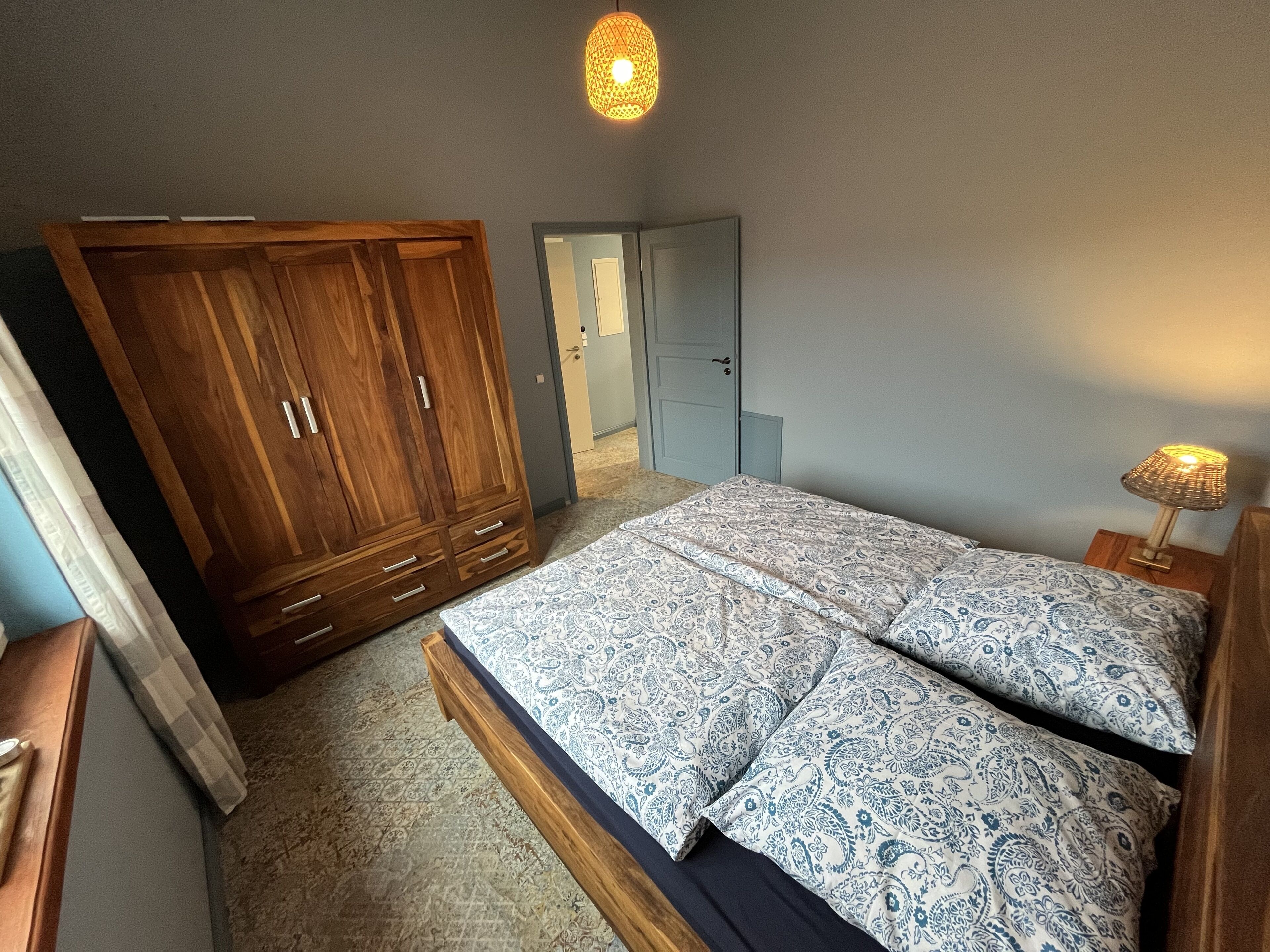 A bedroom with a wooden wardrobe, a bed with patterned bedding, a bedside lamp, and a door leading to another room.