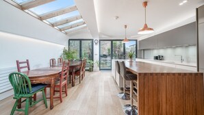 A modern kitchen with a wooden dining table and chairs, a bar area, and large windows allowing natural light. - Beautiful 6bed house near Kensinghton Park (London)
