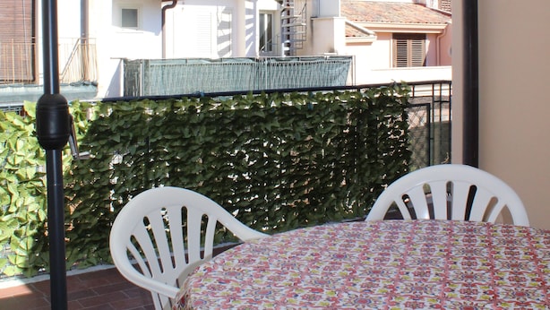 A balcony with a table and chairs, a white umbrella, and a view of buildings in the background.