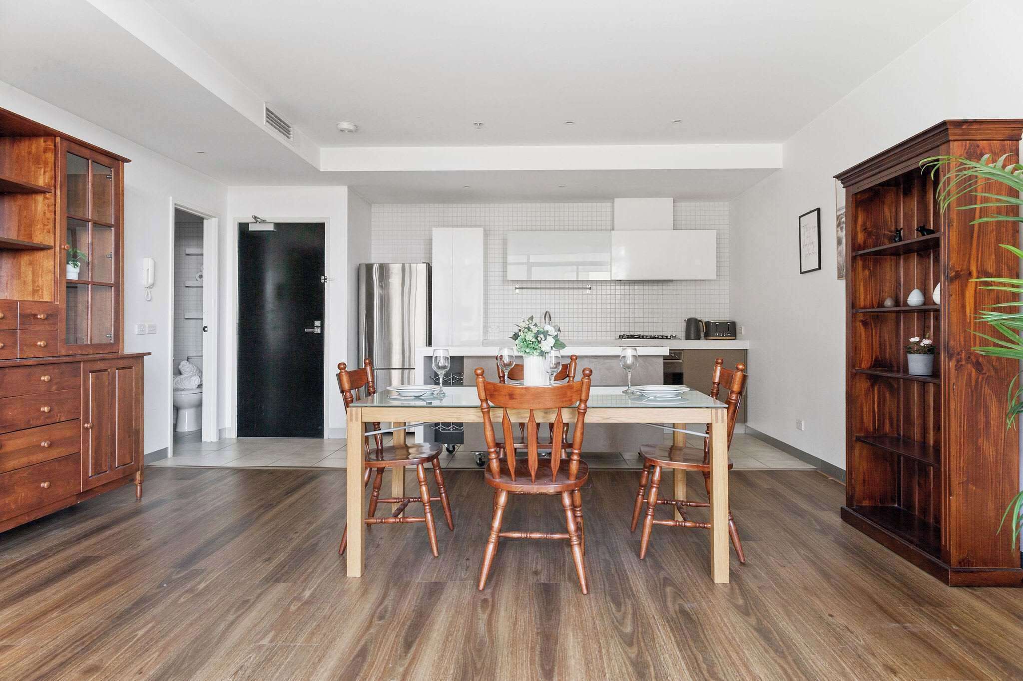 A modern kitchen with white cabinets, a dining table with chairs, and a wooden cabinet with plants.