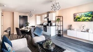 A modern living room with a sofa, a coffee table, and a dining area with chairs and a table. There is a kitchen in the background with cabinets and a microwave. - Spacious & Bright 2 Bed Apartment in Manchester (Manchester)