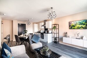 Un salon moderne avec un canapé, une table basse et un coin repas comprenant des chaises et une table. On aperçoit au fond une cuisine équipée de placards et d’un four à micro-ondes.