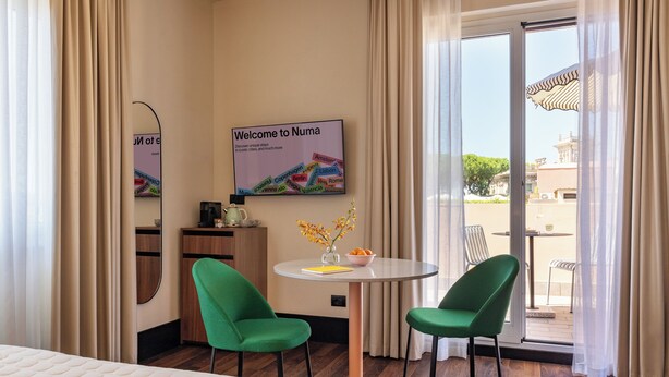 A hotel room with a bed, two green chairs, a small table with a vase and fruit, a coffee maker, and a sign welcoming to Numa. - Numa Rome Barberini (Rome)