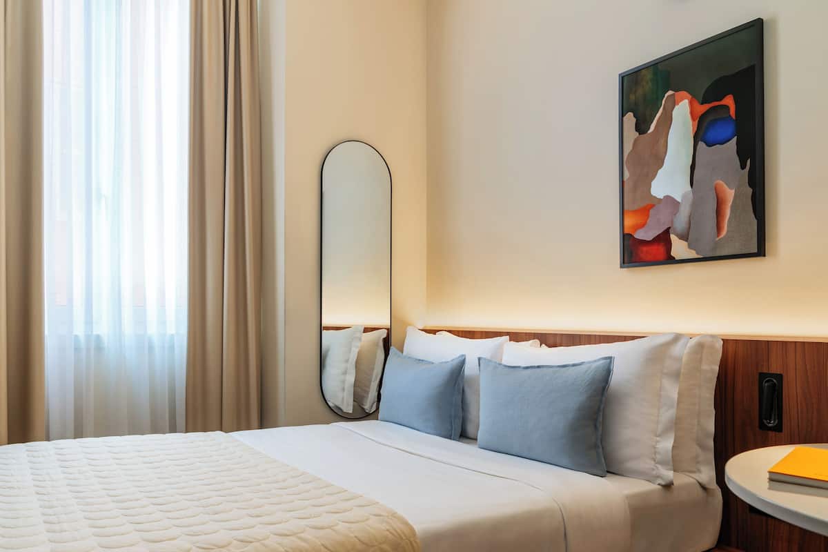A hotel room with a large bed, a round bedside table, a wall-mounted mirror, and a painting.