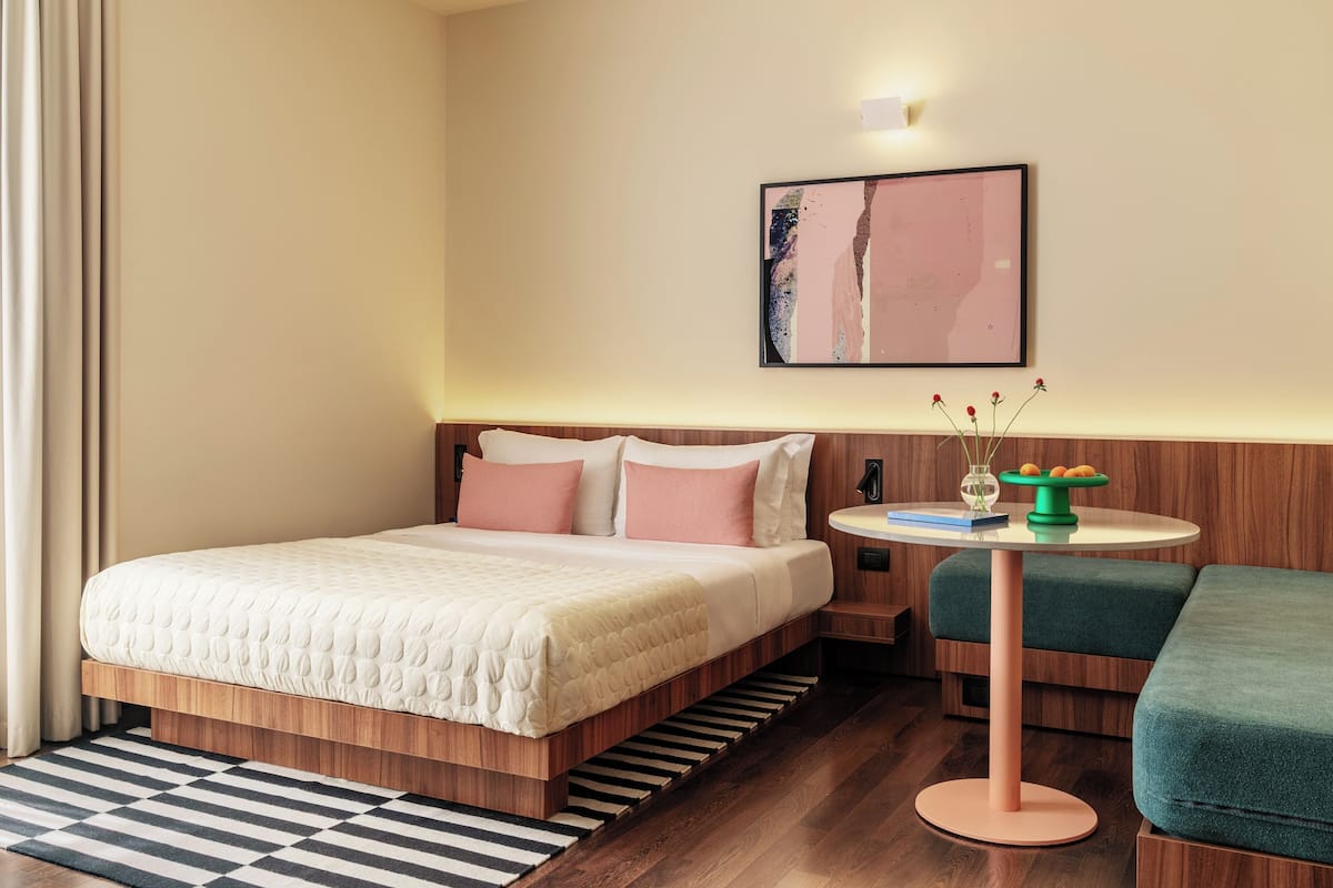 A modern hotel room with a wooden bed, a round table with a vase, and a green chair.