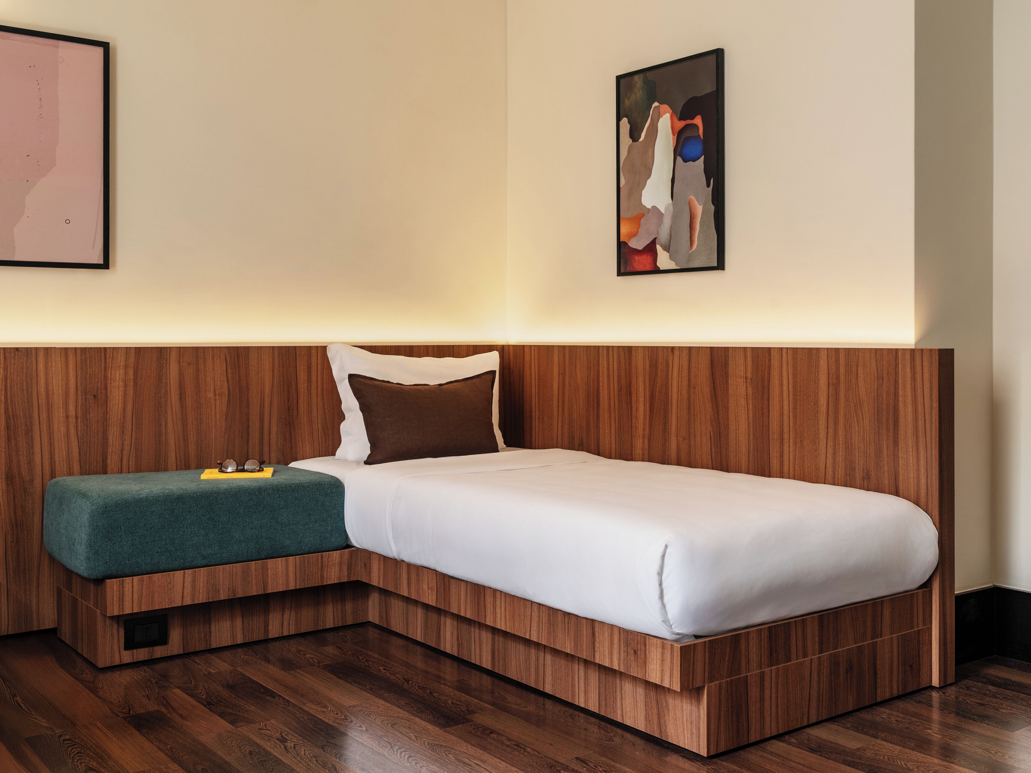 Un hotel moderno con una cama de madera, una sola cama, un pequeño rincón de descanso con un cojín verde y dos obras de arte enmarcadas en las paredes.