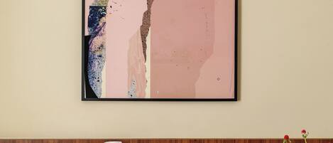 A framed abstract painting on a wall, a bed with a pink pillow, a wooden headboard, and a vase with flowers on a table.