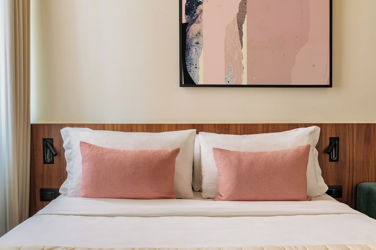 A neatly made bed with a white duvet and two pink pillows, a wooden headboard, and a wall-mounted abstract painting.
