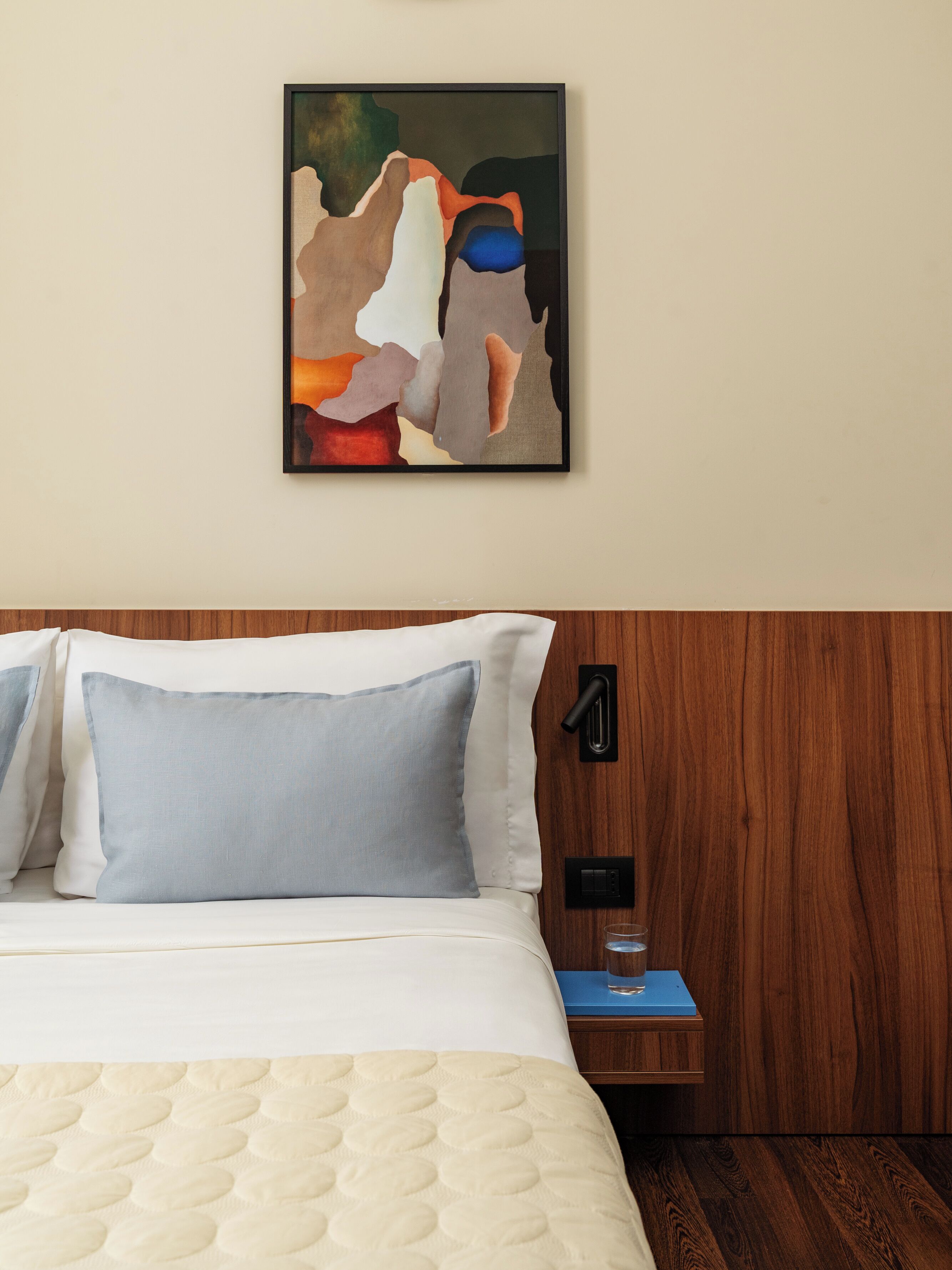 A neatly made bed with a quilted duvet, a bedside table with a glass of water, and a wall-mounted abstract painting.