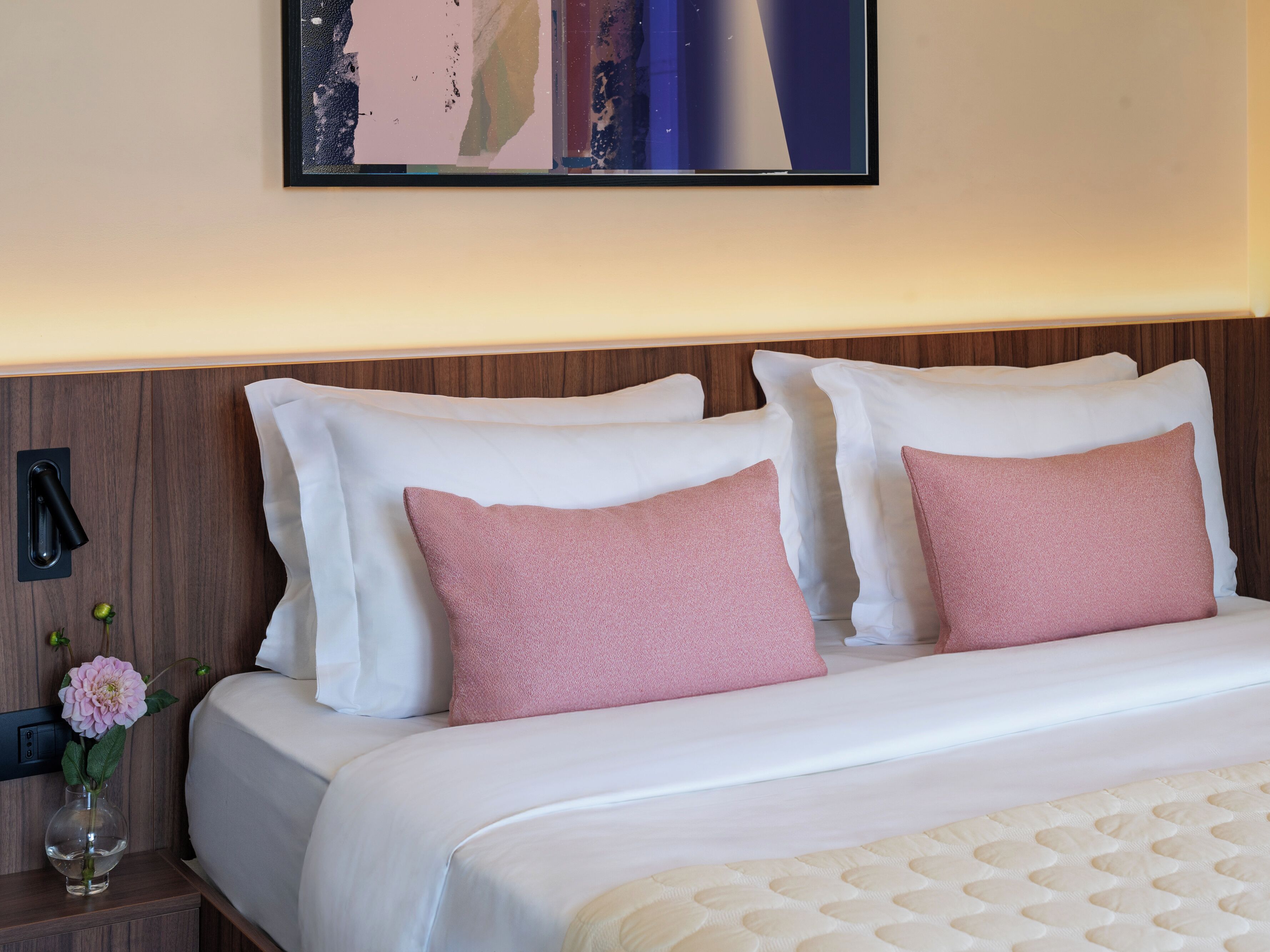 A neatly made bed with white and pink pillows, a wooden headboard, and a wall-mounted artwork above it.