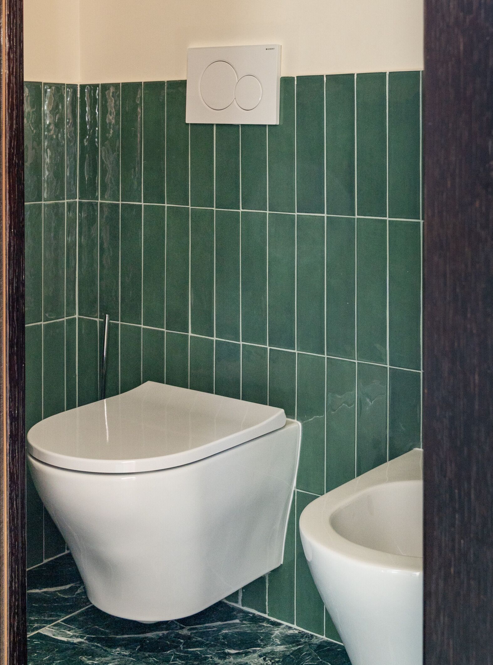 A bathroom with a toilet and a bidet, both in white, against a green tiled wall.