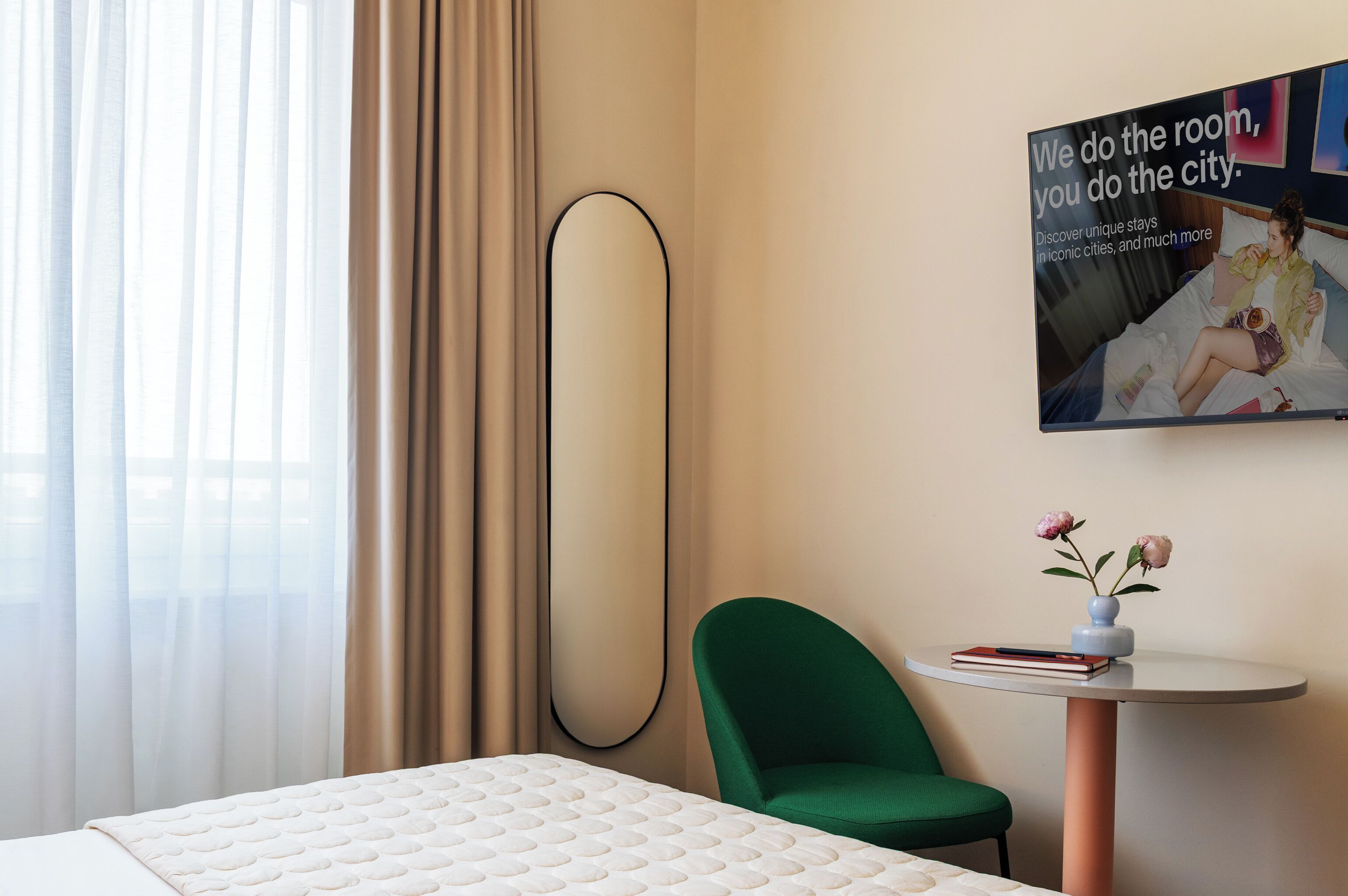 Una habitación de hotel con una cama, una silla verde, una mesita con un florero y un espejo grande, además de un cartel en la pared.